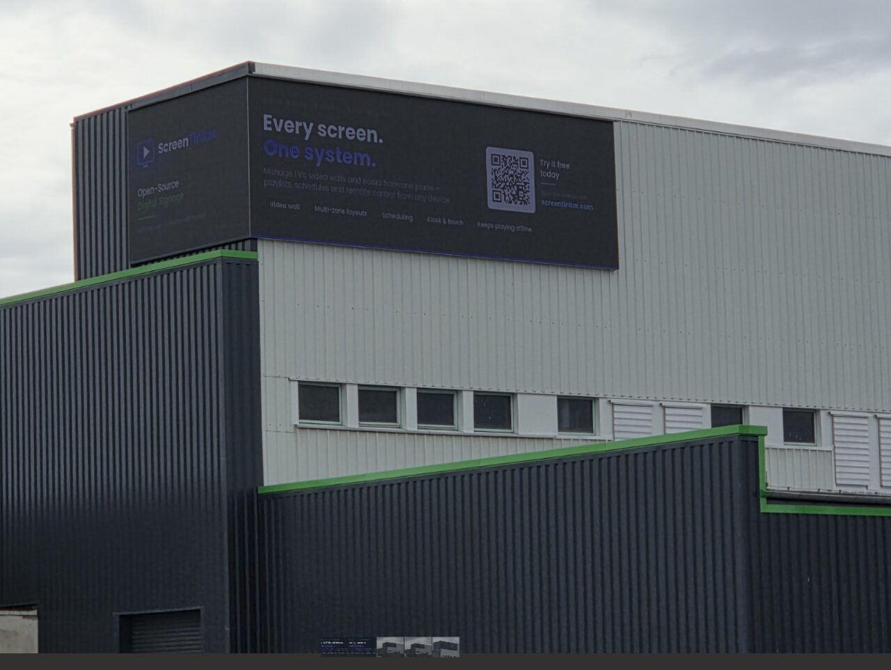 A ScreenTinker advert running on a large outdoor LED video wall mounted on an industrial building